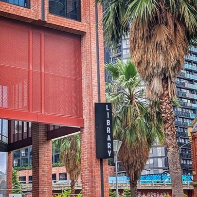 An eye-level shot of a modern red brick building's entrance featuring a large red architectural fabric or mesh screen hanging above an open entryway, a sign with the word LIBRARY on it and city buildings and palm trees in the background.