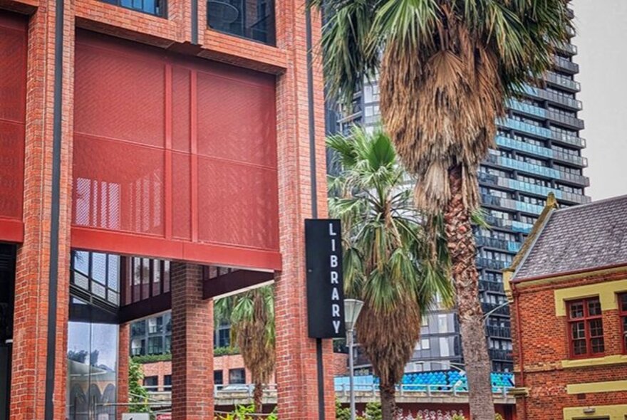 An eye-level shot of a modern red brick building's entrance featuring a large red architectural fabric or mesh screen hanging above an open entryway, a sign with the word LIBRARY on it and city buildings and palm trees in the background.