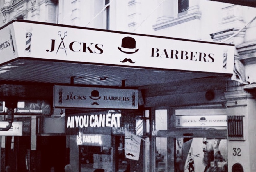 Black and white image of the exterior of Jacks Barbers with lots of signage and a symbol of a bowler hat and moustache.