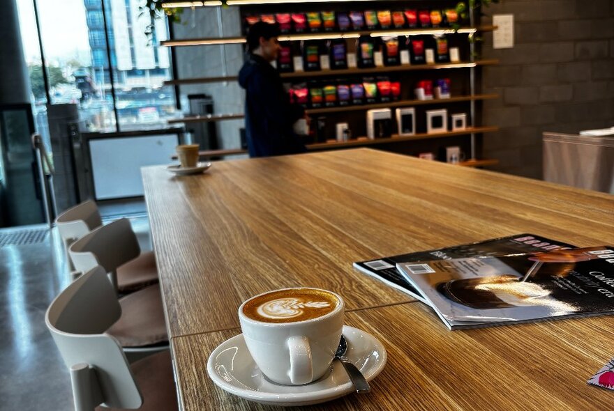 A cup of coffee on a wooden table in a cafe, a shelf in the background with bags of coffee for sale.