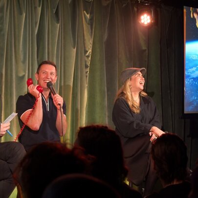 A group of improv comedians seated on stools  on a stage in front of an audience, with a large monitor to their left.