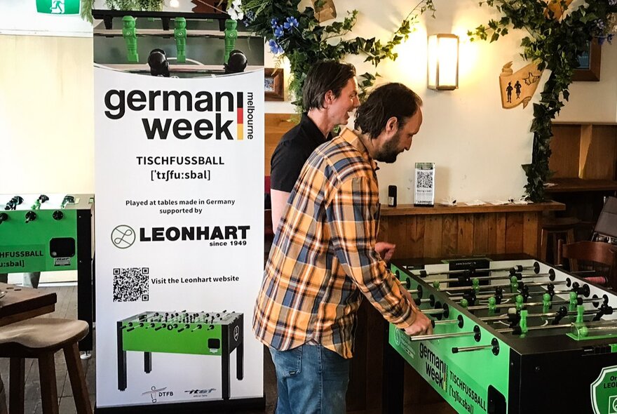 People playing foosball with a German Week poster in the background. 