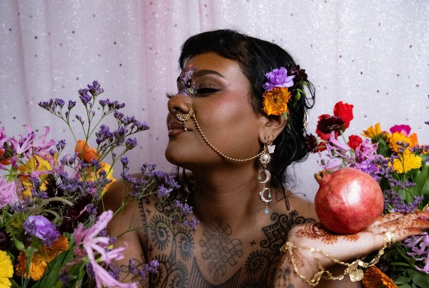 Performer posing with flowers and pomegranate, wearing nose and ear jewellery, her body covered in tattoos.