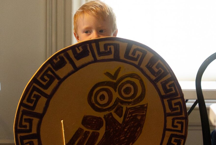A child holding a hand-drawn Greek shield in front of him.