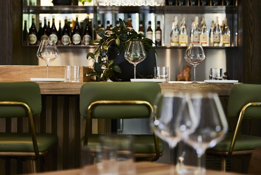 High-backed green stools at a restaurant bar with wine bottles and glasses on display.