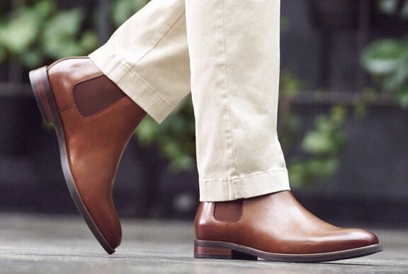 A man walking in a pair of brown leather boots, wearing slim bone-coloured chinos; only his feet and lower legs are seen.