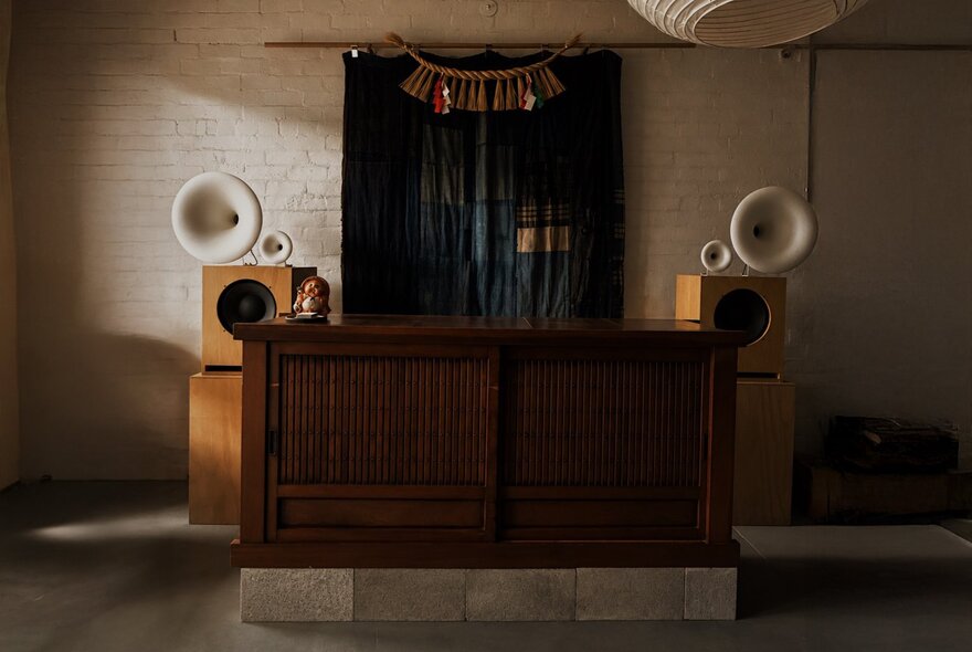 A large wooden counter inside the sparsely  furnished Nordic Japan Gallery space, with two large speakers attached to the white wall.