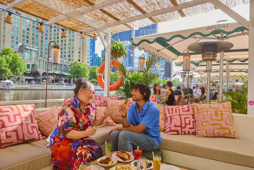 Two friends are sitting on a floating bar under a shady seated area. They are eating snacks and drinking cocktails. 
