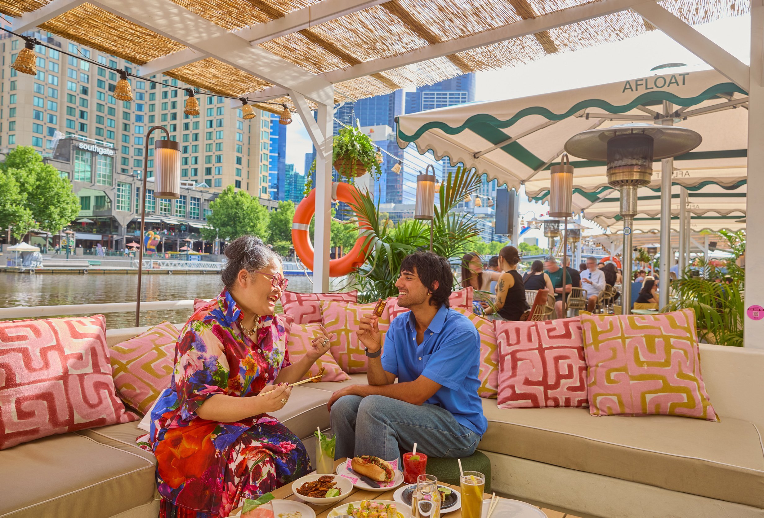 Two friends are sitting on a floating bar under a shady seated area. They are eating snacks and drinking cocktails. 