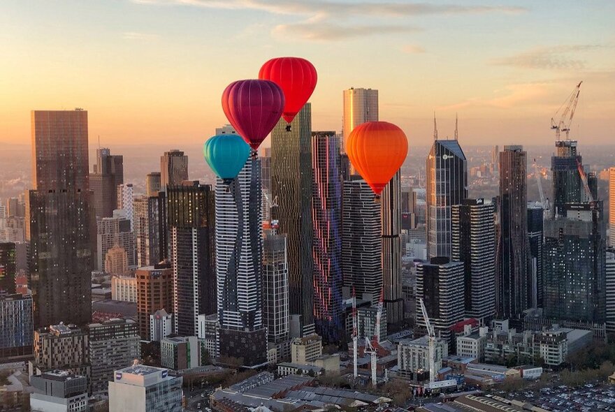 Four hot air balloons floating in front of the Melbourne city skyline during sunrise, with skyscrapers visible in the background.