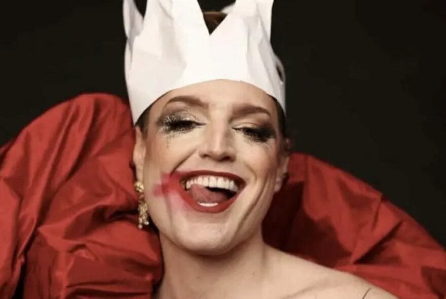Drag artist Reuben Kaye, wearing a paper crown and lipstick, licking his lips.