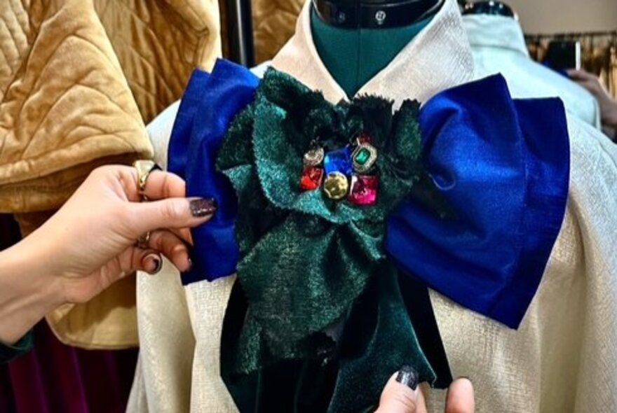 Hands placing a large deep blue and green beaded bow on a the neck of a mannequin's shirt. 