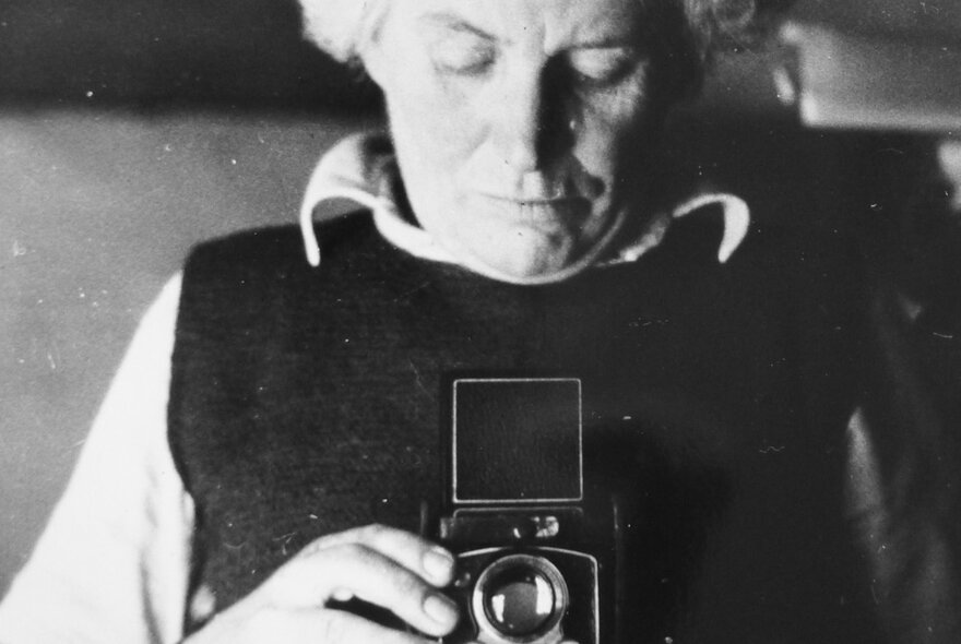 Vintage black and white photograph of Edna Walling looking down into a Kodak box brownie camera she holds in her hands in front of her chest.