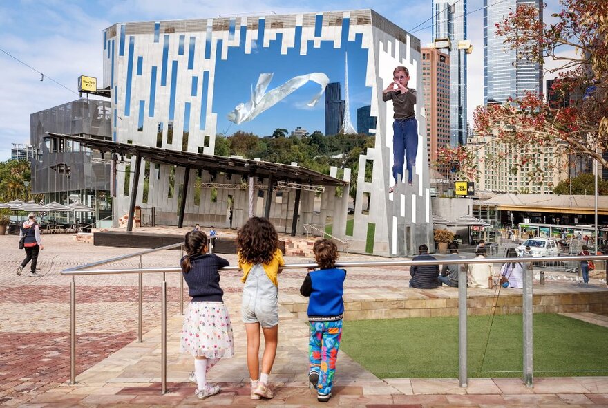 Three children leaning against a railing at Fed Square, looking at the big screen.