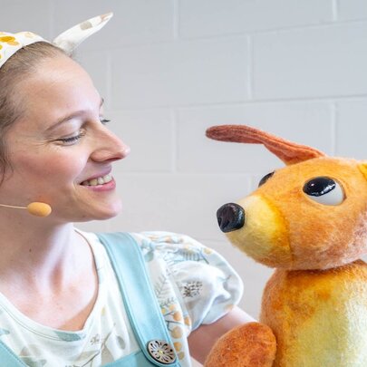 A children's performer in pale blue overalls and a wattle-patterned headband with a cute kangaroo puppet.
