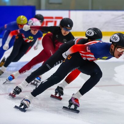 A short-track speed skating race with multiple athletes leaning into a turn on the ice.