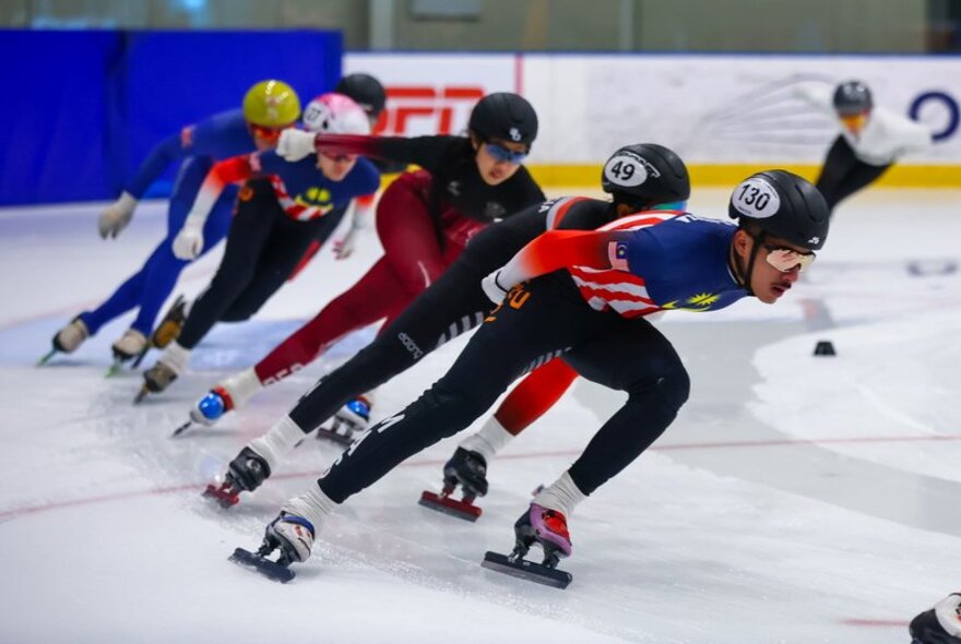 A short-track speed skating race with multiple athletes leaning into a turn on the ice.