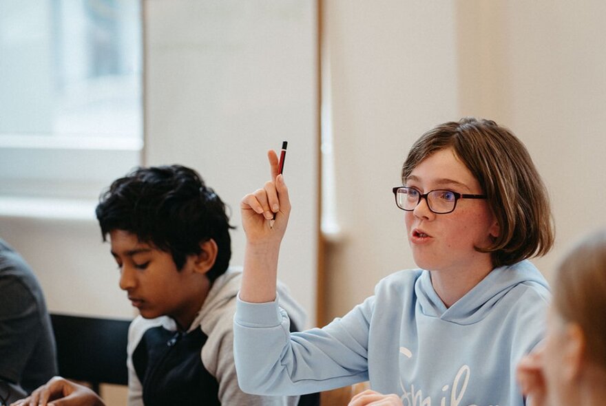 Kids at a class table with one girl raising her hand while holding a pencil. 