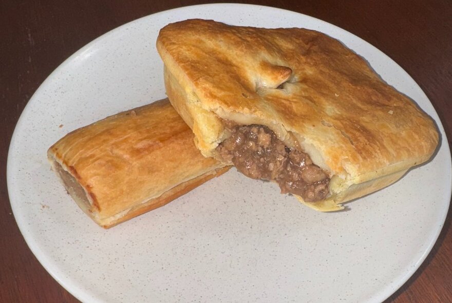 A square meat pie with a bite taken out of it, on a white plate alongside a sausage roll.