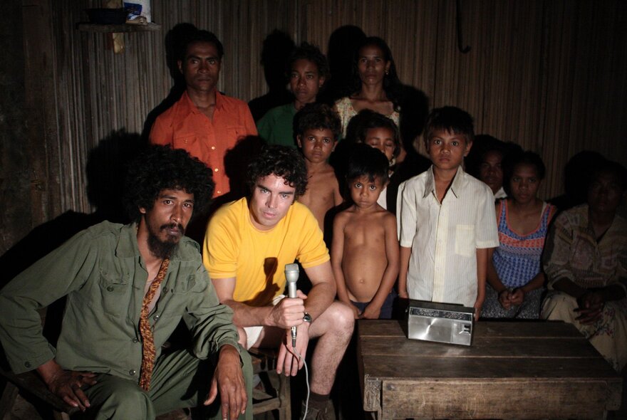 A still from the film Balibo, depicting an East Timorese family inside a room, a Western journalist holding a microphone seated next to them.