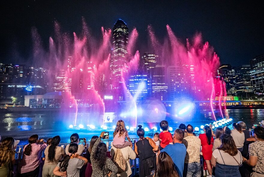 People watch laser lights and water installation illuminating the Yarra River at night.