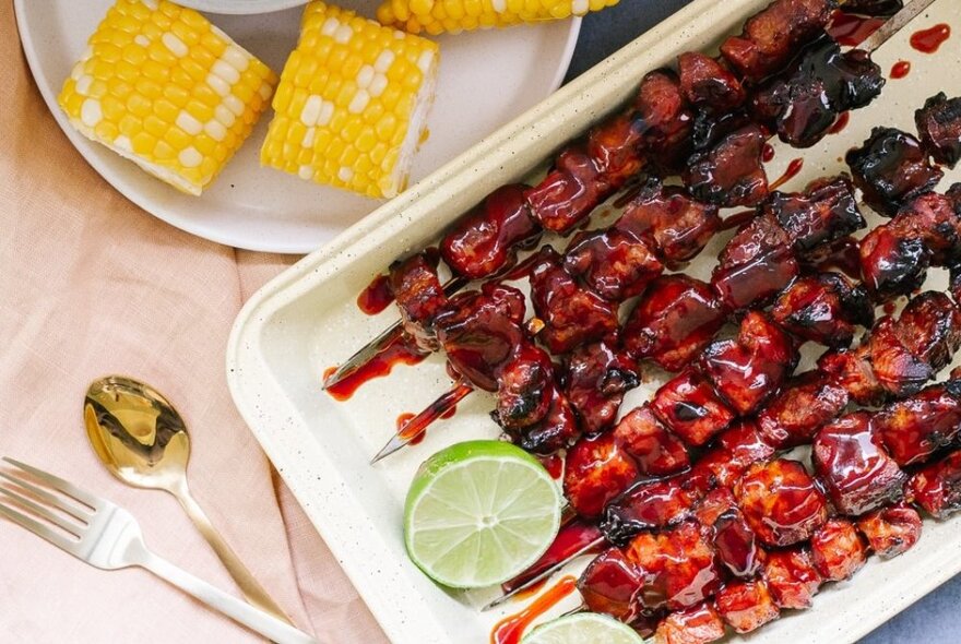 A large dish of red sticky meat on skewers and corn cobs on trays with silver cutlery.