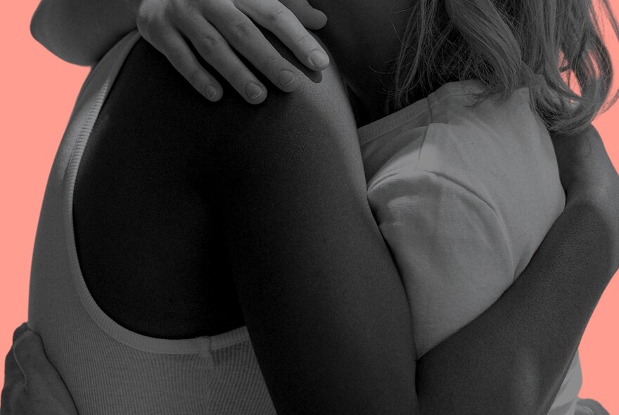 A monochrome close-up from the shoulders down of two people hugging against a peach coloured background. 