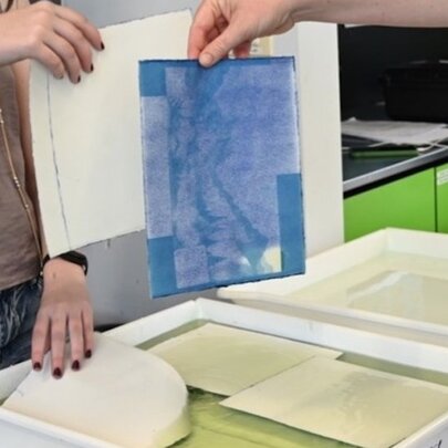 A person rinsing a cyanotype print in a tray of water to reveal a characteristic Prussian blue photographic image.