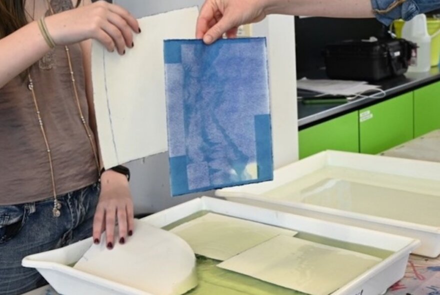 A person rinsing a cyanotype print in a tray of water to reveal a characteristic Prussian blue photographic image.