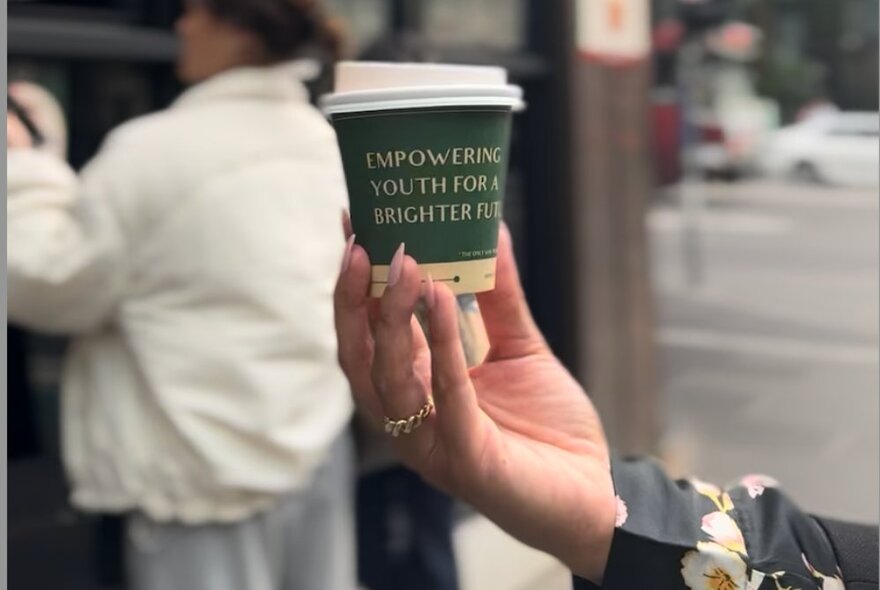 A hand holding up a takeaway coffee cup, a city street and a person in a white jacket behind. 