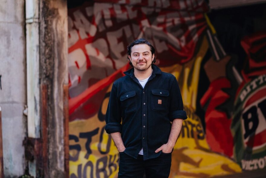 A young man with his hands in his pockets, posing in front of a large yellow and red graffiti mural.