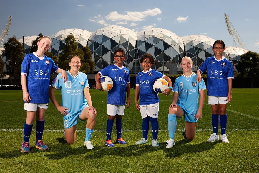 Two players dressed in Melbourne City kit and holding soccer balls in front of AAMI park with four younger kids in dark blue tops.