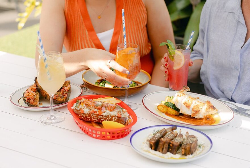 Two people seated at an outside bar table with drinks witih straws and dishes of snack food.