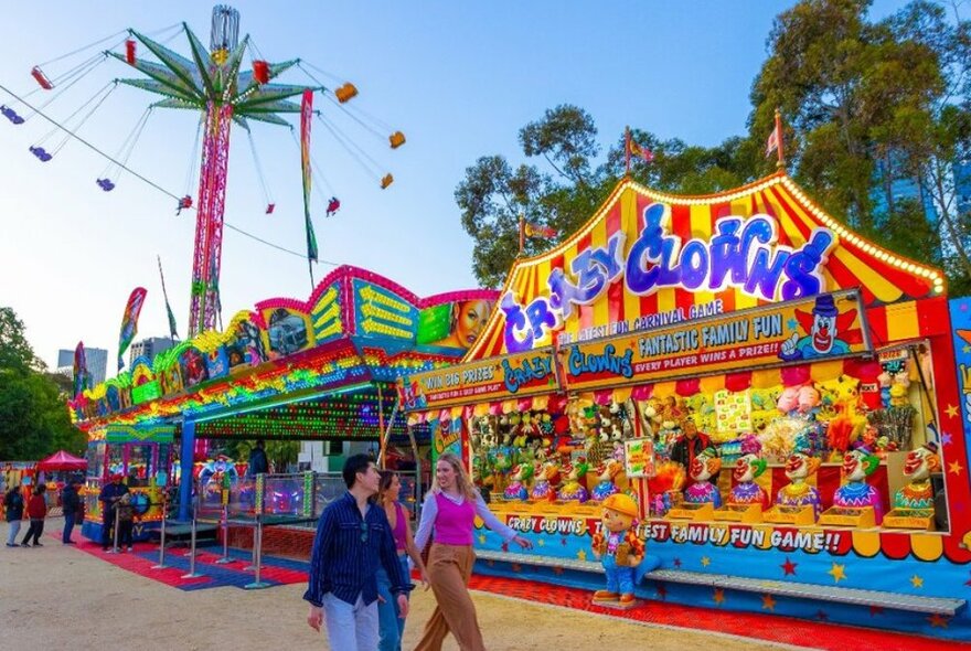 Three friends walking past colourful arcade games and rides at a carnival.