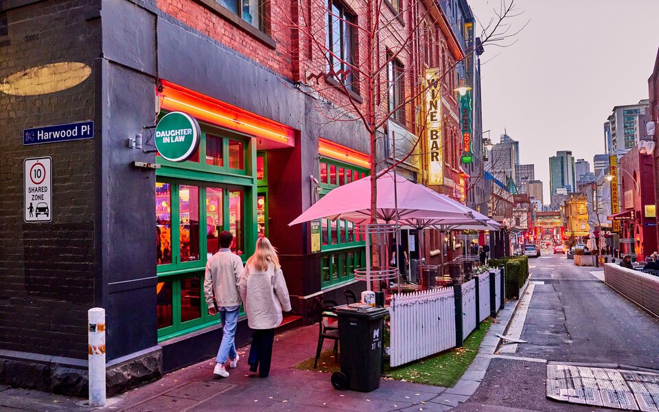 Two people walking down Chinatown outside a restaurant with green window panes and neon lighting.