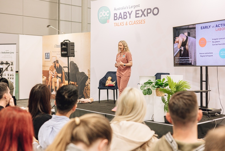 An expert speaker talking on a small stage to a seated audience at The Pregnancy Babies & Children's Expo.