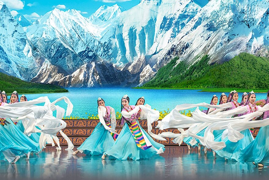Traditional Chinese dancers in long turquoise and white dresses performing on stage in formation with a backdrop depicting snow-covered mountains and lakes.