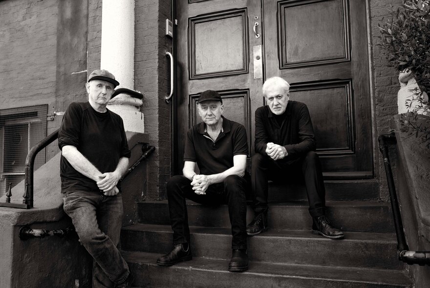 Jazz trio The Necks seated on steps in front of large doors.