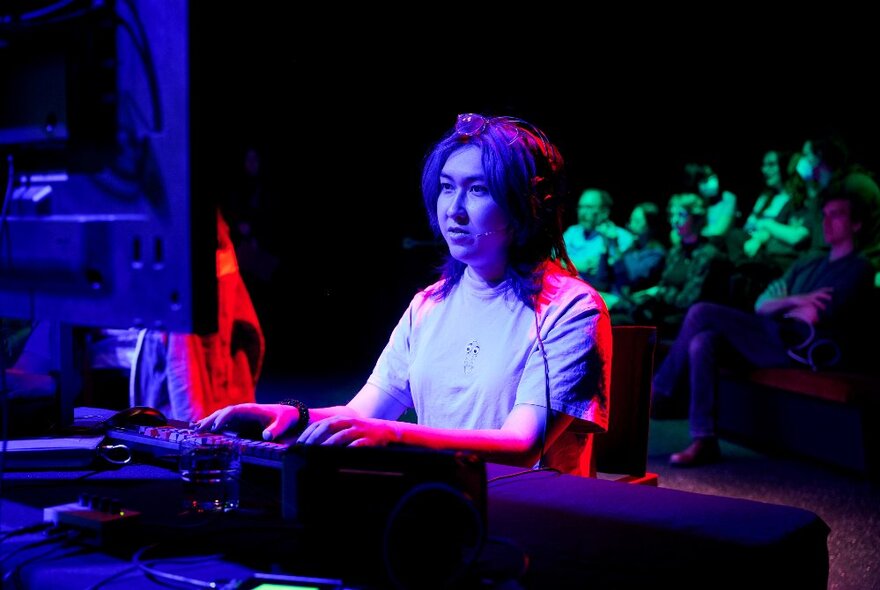 A gamer sitting in front of a screen, competing in a video game competition, with an audience watching behind them.