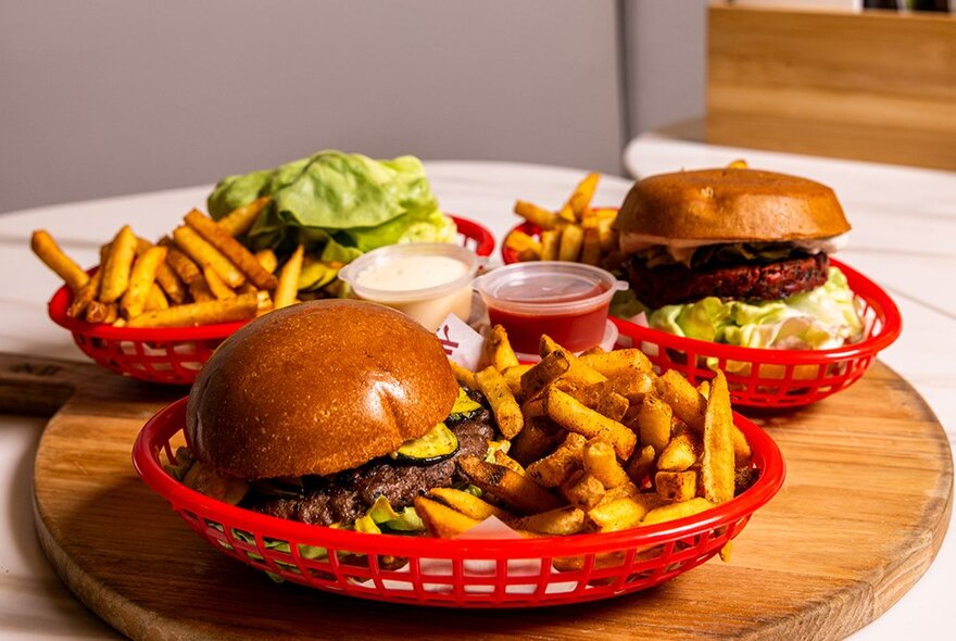 Three classic cheeseburgers with a side of fries and condiments in red baskets, on a round wooden tray, on a restaurant table.