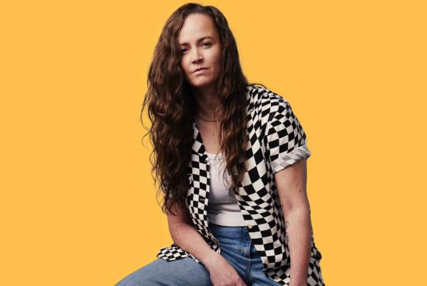 Comedian Zoe Coombs-Marr wearing a black and white chequerboard shirt, long hair flowing down, against a gold background.