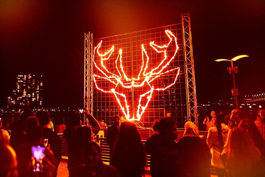 Nighttime at the Firelight Festival with a crowd gathered around an image of a stag's head made of orange glowing light. 