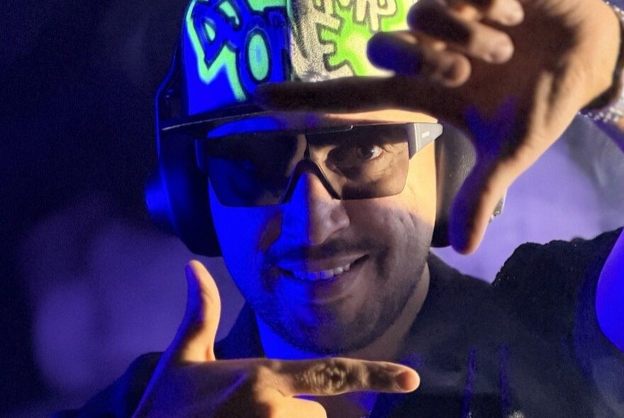 DJ MP ONE with headphones and sunglasses making a square image with both hands in front of his face.