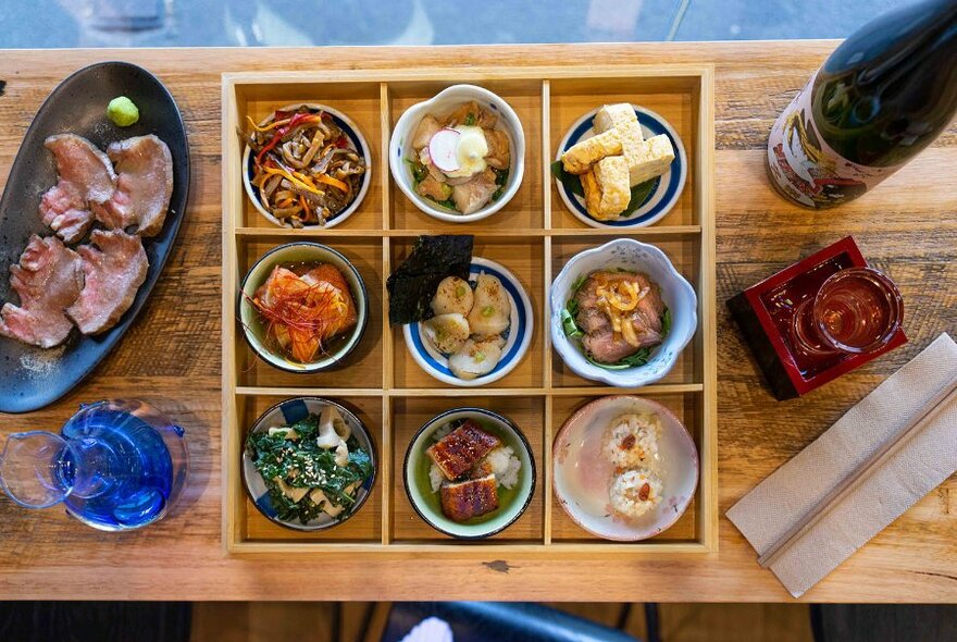 A wooden box with nine compartments filled with Japanese dishes in small bowls.