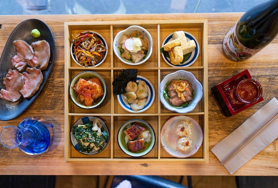 A wooden box with nine compartments filled with Japanese dishes in small bowls.