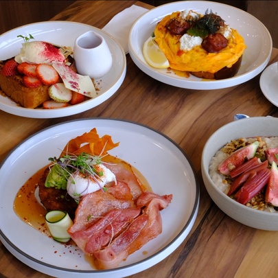 Assorted dishes on a table top including a bacon and poached egg dish, berry French toast, loaded scrambled eggs, a  bowl of porridge and fruits, paired with a café latte.