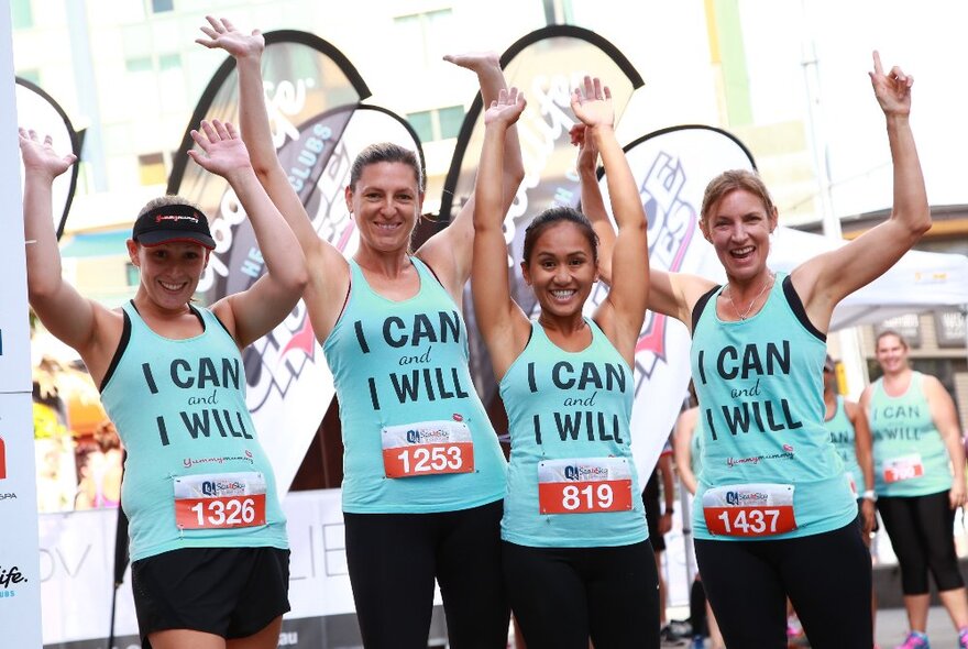 Four charity race participants wearing blue singlets with logos, raising their hands in the air.