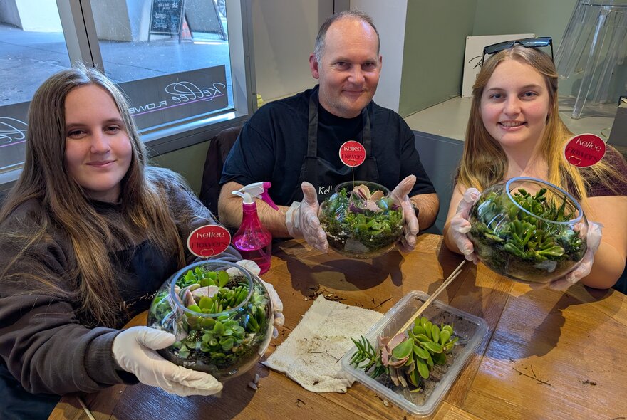 Three people seated at a table wearing work gloves, holding glass terrariums.