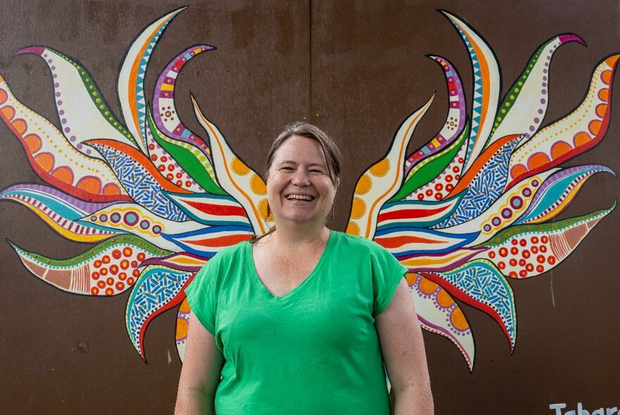 A smiling woman in a green top lines up perfectly with painted coloured wings on the wall behind. 