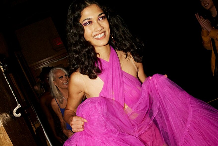 A smiling fashion model wearing a bright fuschia pink gown and walking backstage at a fashion show.